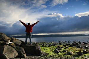 man-on-rocks-beach-arms-raised-to-sky-clouds